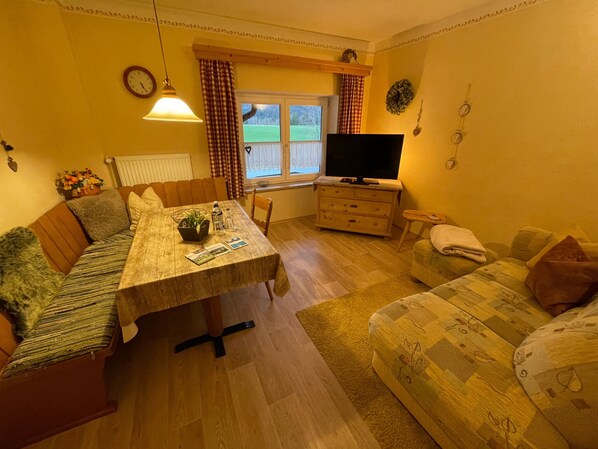 A cozy living room with a wooden dining table, a sofa, a TV, and a window with curtains.