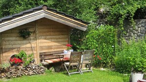 A wooden cabin with a table and chairs outside, surrounded by a garden with plants and a stone wall.