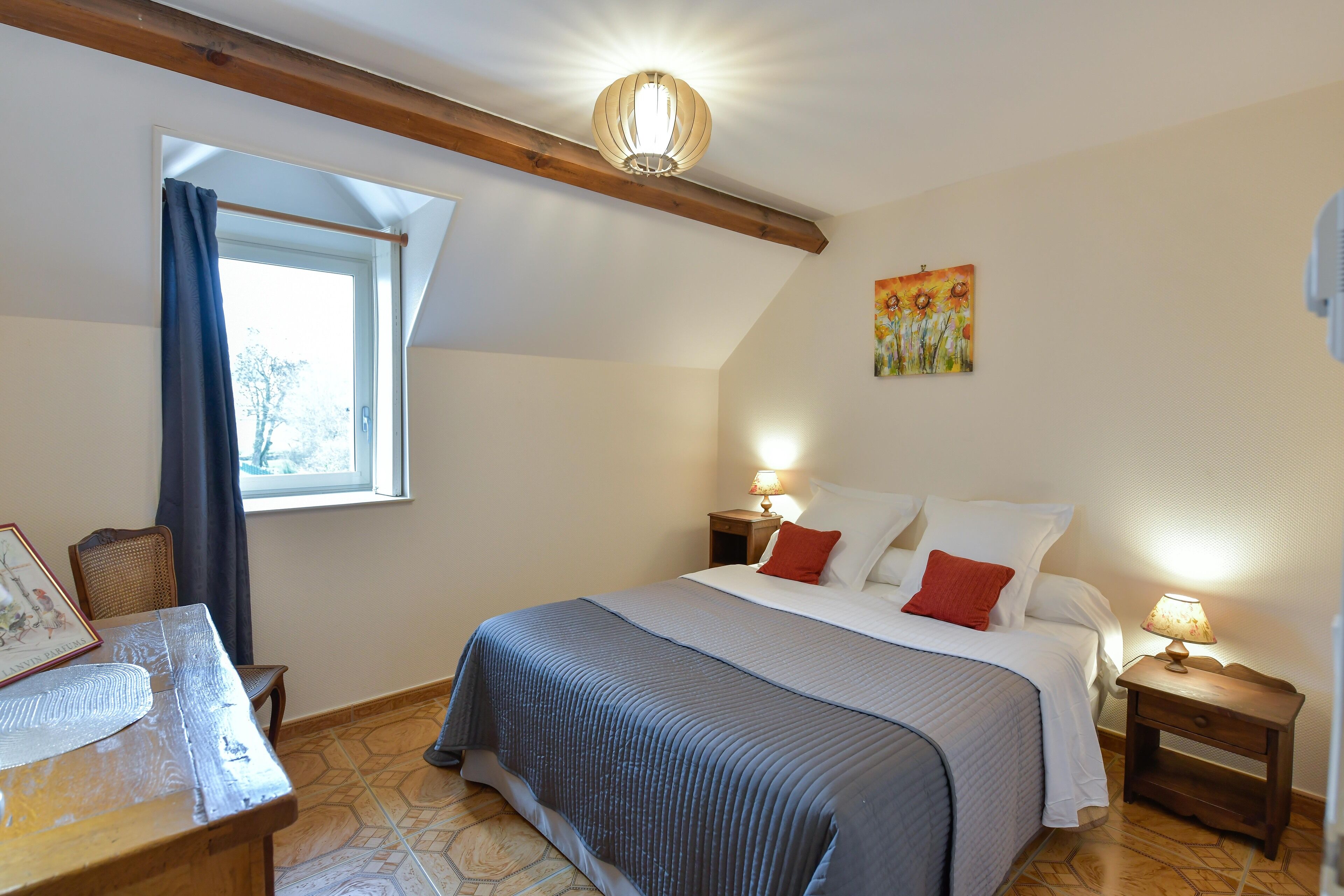 Une chambre à coucher avec un lit, des tables de chevet, une fenêtre avec des rideaux, un tableau au mur et une poutre en bois au plafond.