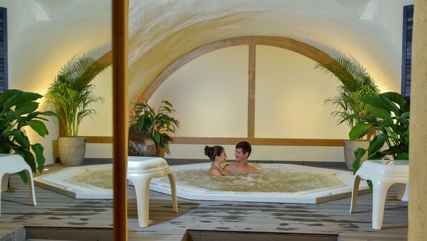 A couple in a hot tub with a wooden arch ceiling and potted plants. - Le clos champagnac at Hotel Golf Chateau de Chailly (Chailly-sur-Armancon)