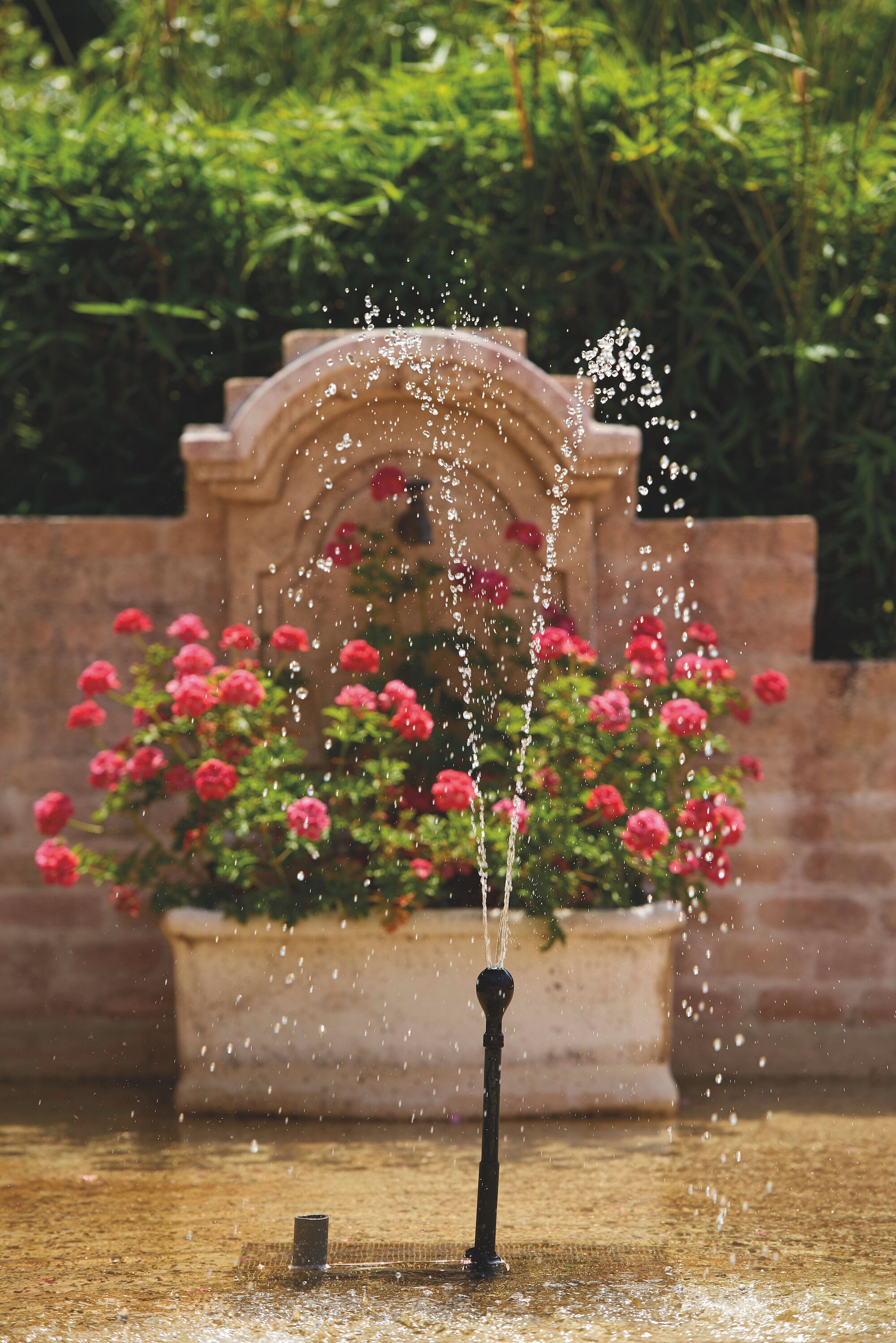 A fountain with water spouting upwards, surrounded by a planter with pink flowers, set against a brick wall and greenery.
