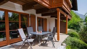 An outdoor patio with a table and chairs, large windows, and a wooden balcony. - Apartment 2/1294, 70sqm, terrace, kitchenette, 2 bedrooms, 1 living room, 5 p., WLAN (Ruhpolding)
