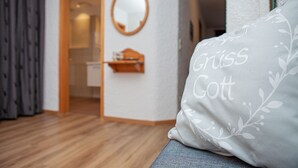 A room with a wooden floor, a grey sofa with a pillow labeled "Griss Gott," a wooden door, and a wall-mounted mirror. - Apartment 2/1294, 70sqm, terrace, kitchenette, 2 bedrooms, 1 living room, 5 p., WLAN (Ruhpolding)