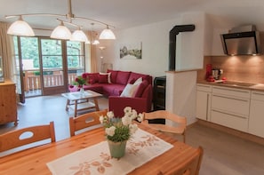 Uma sala de estar moderna com sofá vermelho, mesa de jantar de madeira e área de cozinha com armários brancos e coifa de aço inoxidável.