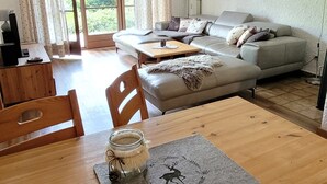 A cozy living room with a sofa, a wooden dining table, and a view of the outdoors through large windows. - Apartment 2/1294, 70sqm, terrace, kitchenette, 2 bedrooms, 1 living room, 5 p., WLAN (Ruhpolding)