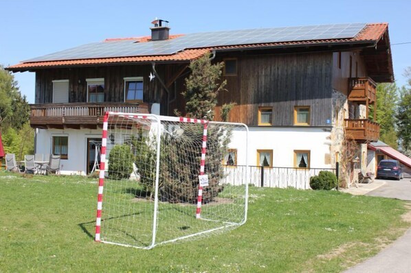 A rustic wooden house with a solar panel roof, a fenced soccer goal, and a balcony. - Ferienwohnung I.OG, 50m², 2-4 Pers., Du/WC, Ostbalkon (Teisendorf)