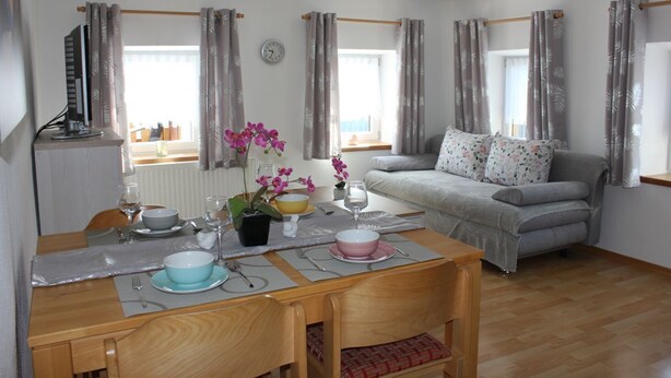 Una sala de estar con una mesa de comedor lista para dos personas, un sofá gris con cojines estampados y una ventana con cortinas.