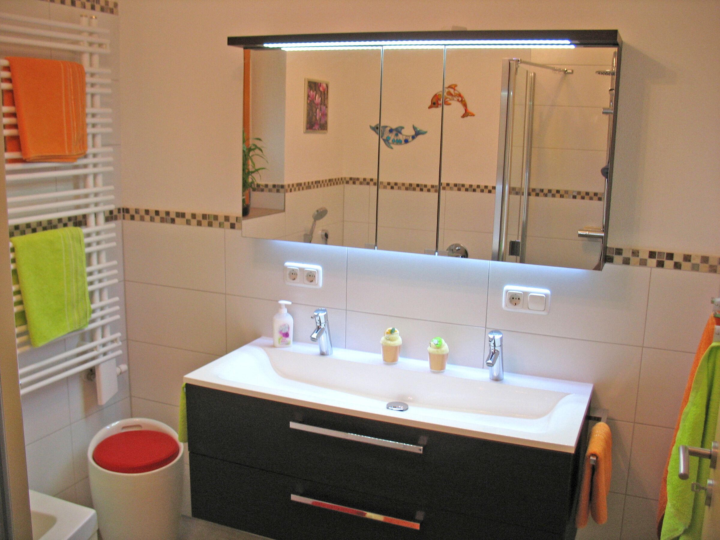 Een badkamer met een wit wastafel, twee spiegels en een douche. Handdoeken liggen op een rek en de toiletbril is rood.