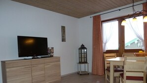 A living room with a flat-screen TV on a wooden cabinet, a table with chairs, a small table lamp, and a window with curtains. - Two-room apartment, 40m², bedroom and living room, kitchenette, balcony (Reit im Winkl)