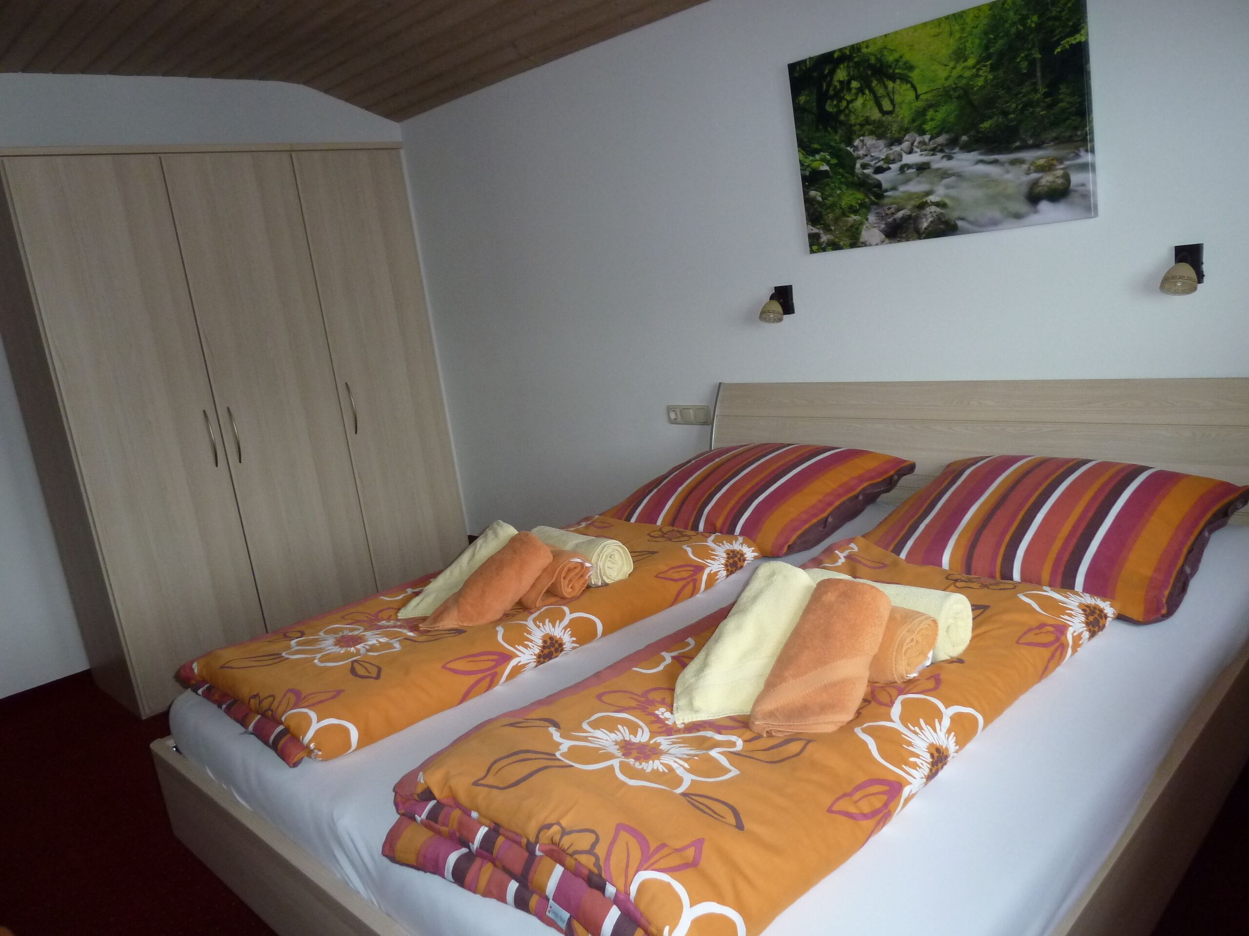 Uma cama de casal com roupa de cama floral e listrada, um guarda-roupa de madeira e um quadro de um riacho em uma floresta na parede.