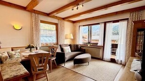 A cozy living room with a wooden ceiling, a sofa, a dining table with chairs, and large windows with curtains. - Drei-Raum-Ferienwohnung (7) Unterberg, 82 qm, Bad/WC, Extra-Schlafzimmer, Küche, Balkon (Reit im Winkl)