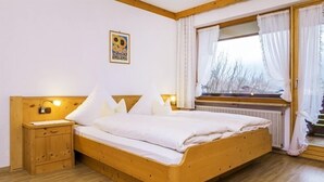 A wooden bed with white bedding in a room with a wooden floor, a nightstand, and a window with curtains. - Drei-Raum-Ferienwohnung (7) Unterberg, 82 qm, Bad/WC, Extra-Schlafzimmer, Küche, Balkon (Reit im Winkl)