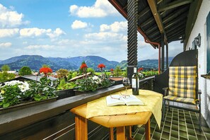 A wooden table with a book and wine glass, a checkered chair, and a view of mountains and greenery. - Zwei-Raum-Ferienwohnung "Bergblick", 37m², extra Schlafzimmer, sep. Küche und Balkon (Reit im Winkl)