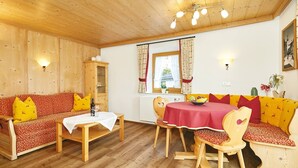 A cozy room with wooden walls and ceiling, a sofa with red and yellow cushions, a dining table with a red tablecloth, and a window with curtains. - Zwei-Raum-Ferienwohnung "Bergblick", 37m², extra Schlafzimmer, sep. Küche und Balkon (Reit im Winkl)