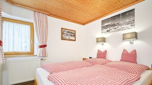 A bedroom with a wooden ceiling, a bed with red checkered bedding, a window with white curtains, and a wall-mounted picture. - Zwei-Raum-Ferienwohnung "Bergblick", 37m², extra Schlafzimmer, sep. Küche und Balkon (Reit im Winkl)