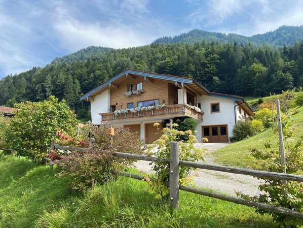 Une maison en bois à deux étages, dotée d’un balcon, entourée d’une végétation luxuriante et avec des montagnes en arrière-plan.