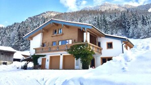 Ein zweigeschossiges Holzhaus mit Balkon, umgeben von schneebedeckten Bäumen und Bergen.