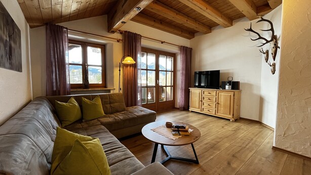 En hyggelig stue med træloft, en sofa med puder, et sofabord med bøger, et fjernsyn på et træskab og et vindue med gardiner.