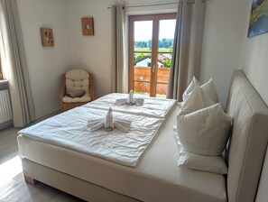 A neatly made bed with white linens and pillows, a wicker chair, and a view of a wooden deck through a glass door.