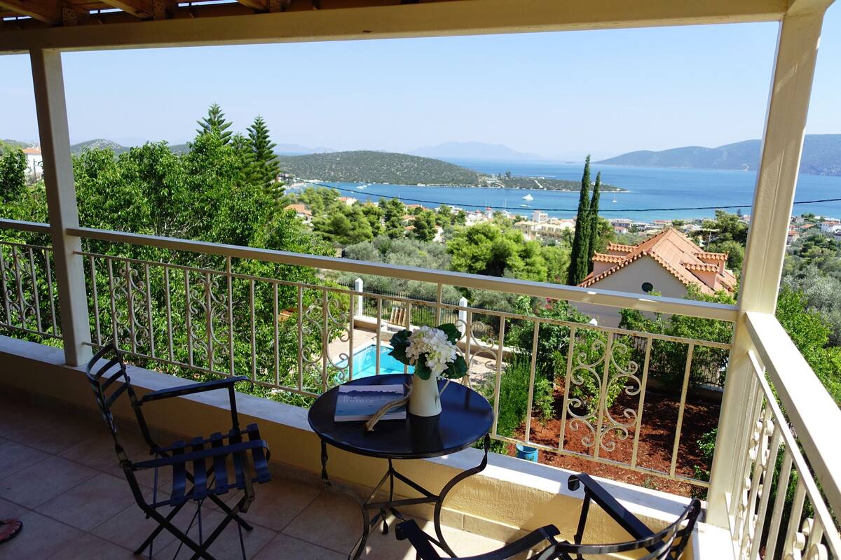 Un balcón con mesa y sillas, con vistas a un paisaje pintoresco de un pueblo costero y el mar.