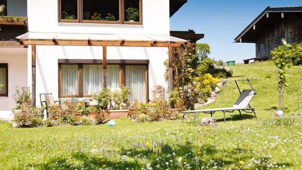 A modern house with a white exterior, large windows, and a well-maintained garden with various plants and flowers. - Vacation apartment 36 sqm, no. 1, covered terrace, kitchen, 1 living/bedroom, WLAN (Ruhpolding)