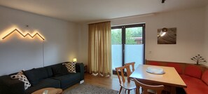 A modern living room with a sofa, a dining table with chairs, a large window with curtains, and a wall-mounted light fixture. - Bungalow 125sqm, terrace, garden, kitchen, 3 bedrooms, living room, 2 bathrooms, WLAN (Ruhpolding)