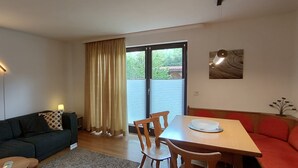 Una sala de estar moderna con un sofá, una mesa de comedor y sillas. Hay un ventanal con cortinas, una lámpara de techo y una mesita con una lámpara.