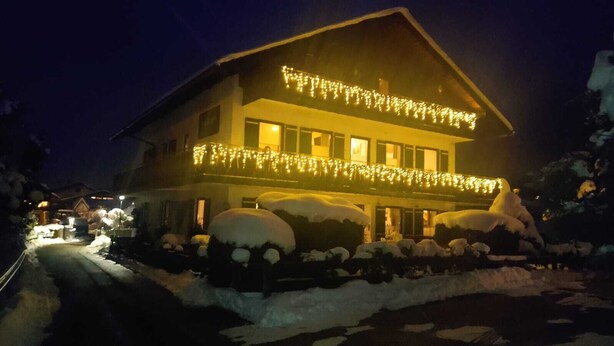 Un edificio de dos plantas con un balcón, arbustos cubiertos de nieve y luces colgantes.