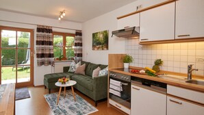 A modern kitchen with white cabinets, a built-in oven, and a dishwasher. There is a green sofa and a small table with fruit and a glass of wine. - Ferienwohnung "Rosenduft", 58 qm, Erdgeschoss, 1 separates Schlafzimmer, Südseite mit Terrasse (Höslwang)