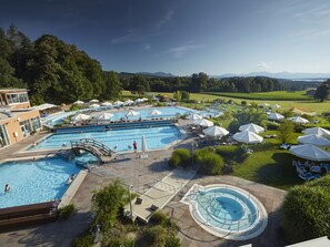 A large outdoor swimming pool with multiple lanes, surrounded by lounge chairs and umbrellas, set against a backdrop of trees and distant mountains.