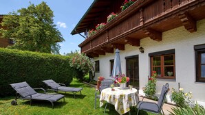 A traditional chalet with a balcony, a garden with lounge chairs, and a table with a floral tablecloth. - Ferienwohnung "Rosenduft", 58 qm, Erdgeschoss, 1 separates Schlafzimmer, Südseite mit Terrasse (Höslwang)