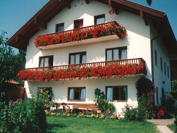 A two-story building with balconies adorned with red flowers, multiple windows, and a garden area with various plants and flowers.