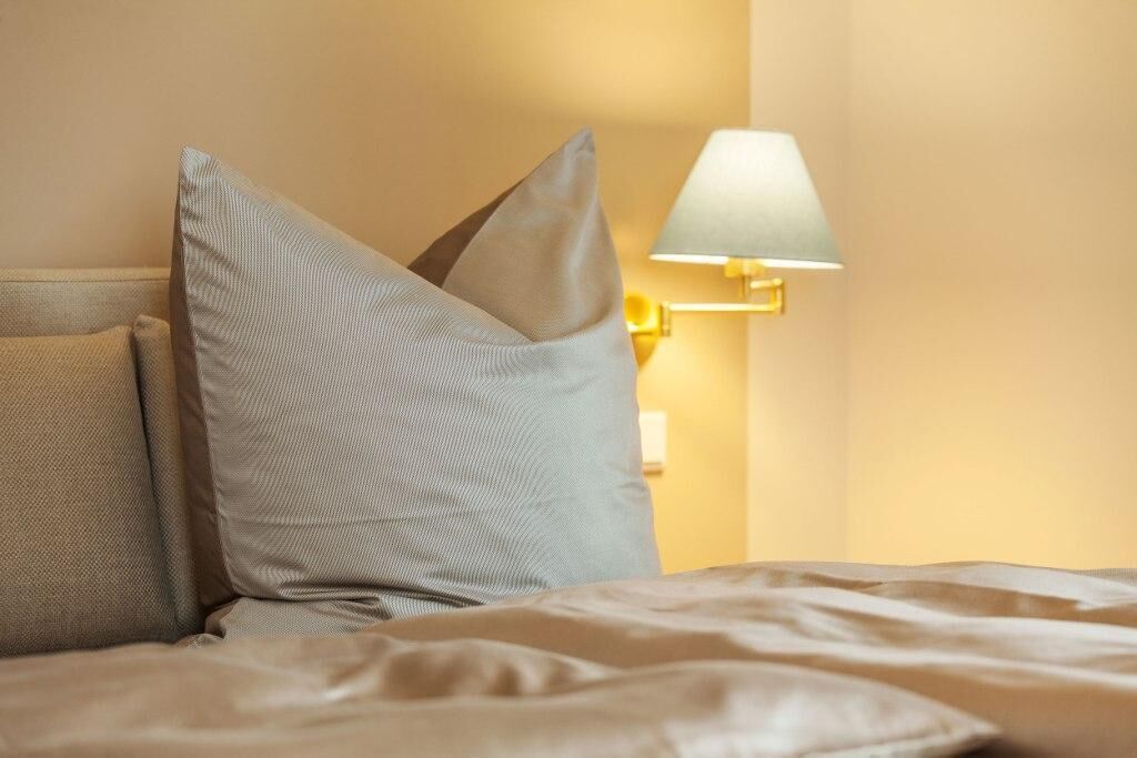 A bed with beige bedding and two pillows, a wall-mounted lamp, and a plain wall.