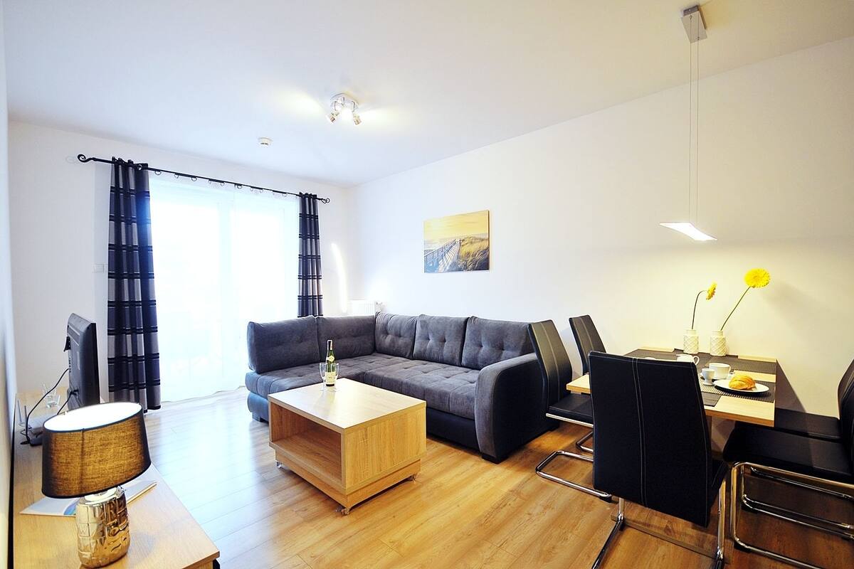 Ein modernes Wohnzimmer mit einem grauen Ecksofa, einem Holztisch, einem Esstisch mit Stühlen und einer Lampe.