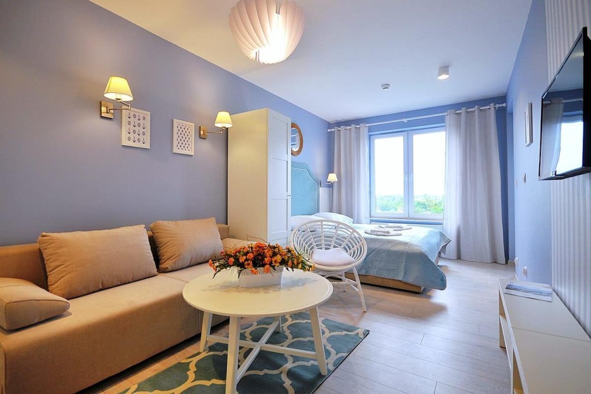 Ein modernes Hotelzimmer mit einer Couch, einem kleinen runden Tisch mit Blumenvase, einem Bett mit blauer Bettdecke und einem großen Fenster mit Vorhängen.