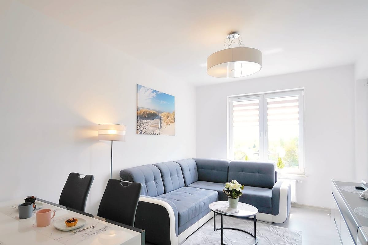 Ein modernes Wohnzimmer mit einem blauen Ecksofa, einem Esstisch mit Stühlen, einer Stehlampe und einem großen Fenster mit Jalousien.