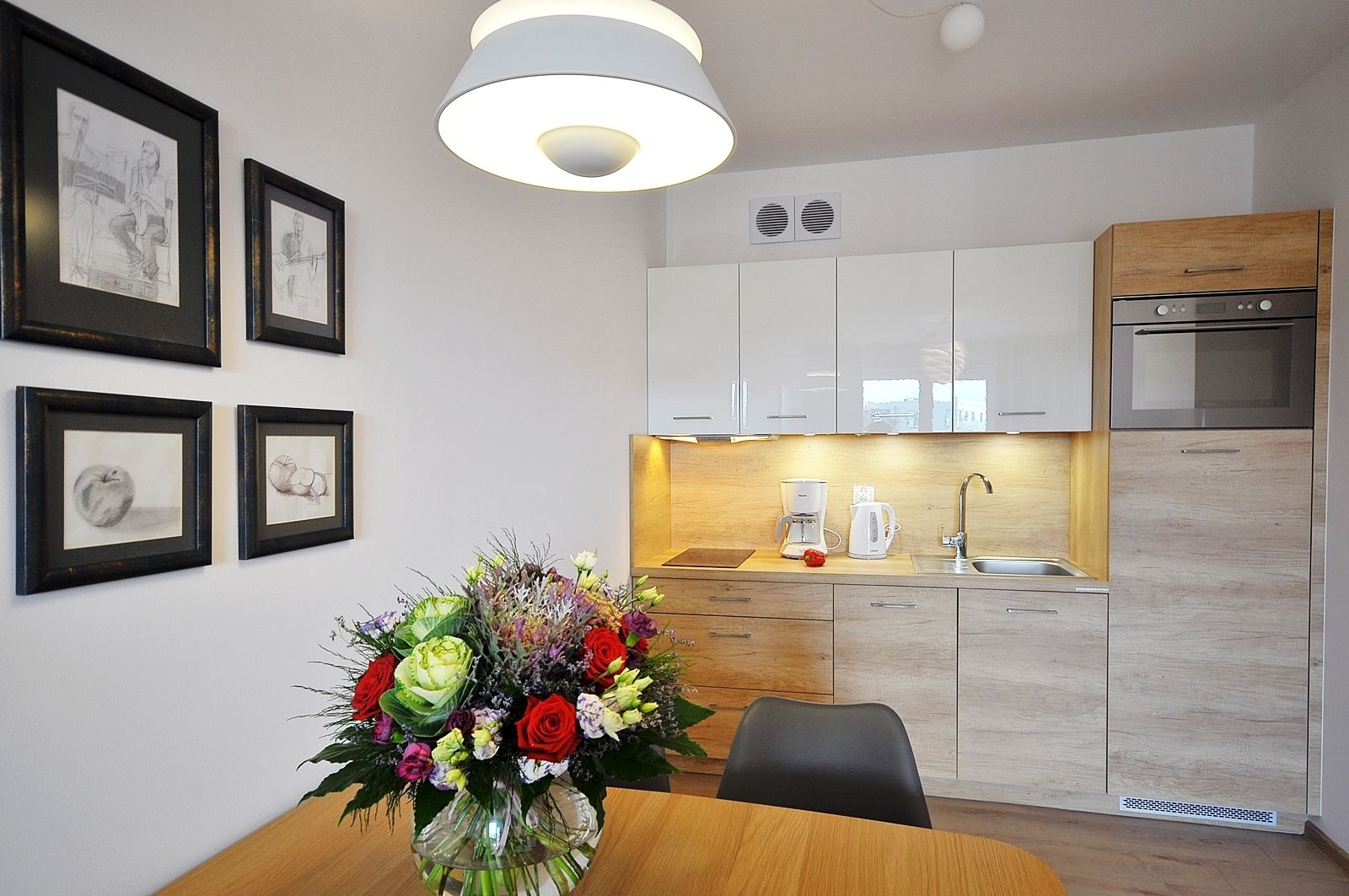 A modern kitchen with white cabinets, a built-in oven, and a coffee maker. There are framed artworks on the wall and a vase of flowers on the table.