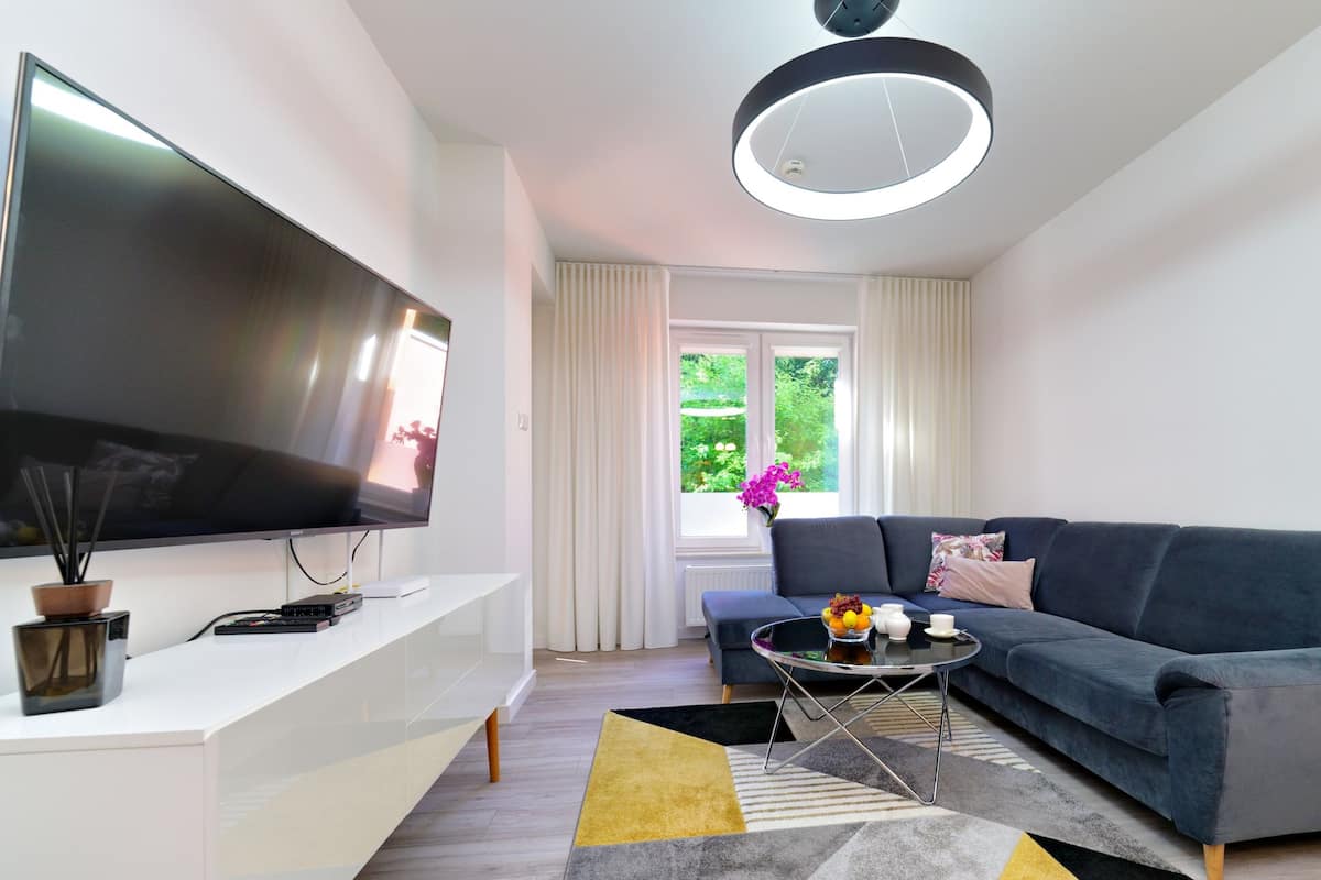 Ein modernes Wohnzimmer mit einem Flachbildfernseher, einem dunkelblauen Sofa, einem Couchtisch und einem großen Fenster mit Vorhängen.