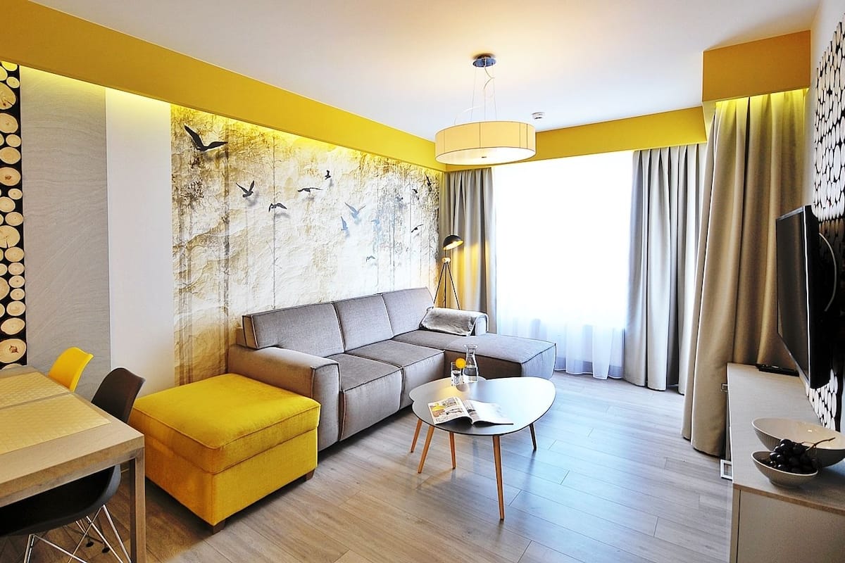 Ein modernes Wohnzimmer mit einer Couch, Hocker und Couchtisch. Ein Esstisch mit einer Champagnerflasche steht ebenfalls im Raum. Ein großes Fenster mit Vorhängen sorgt für Licht.