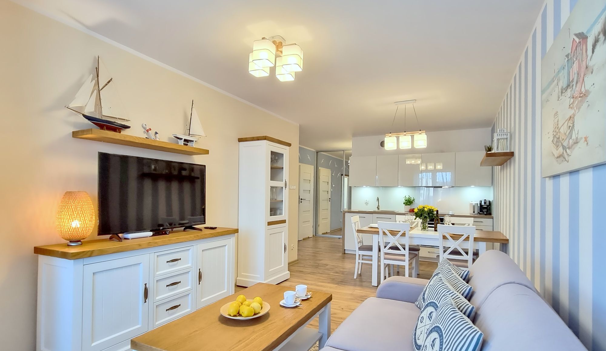 A modern living room with a flat-screen TV, wooden cabinets, a dining area with a table and chairs, and a kitchen in the background.