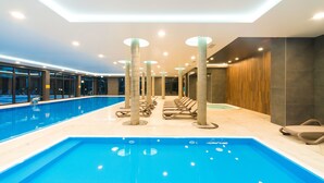 Une piscine intérieure moderne, dotée d’une eau cristalline, entourée de fauteuils et de chaises longues, et dont le plafond est agrémenté d’un éclairage encastré.