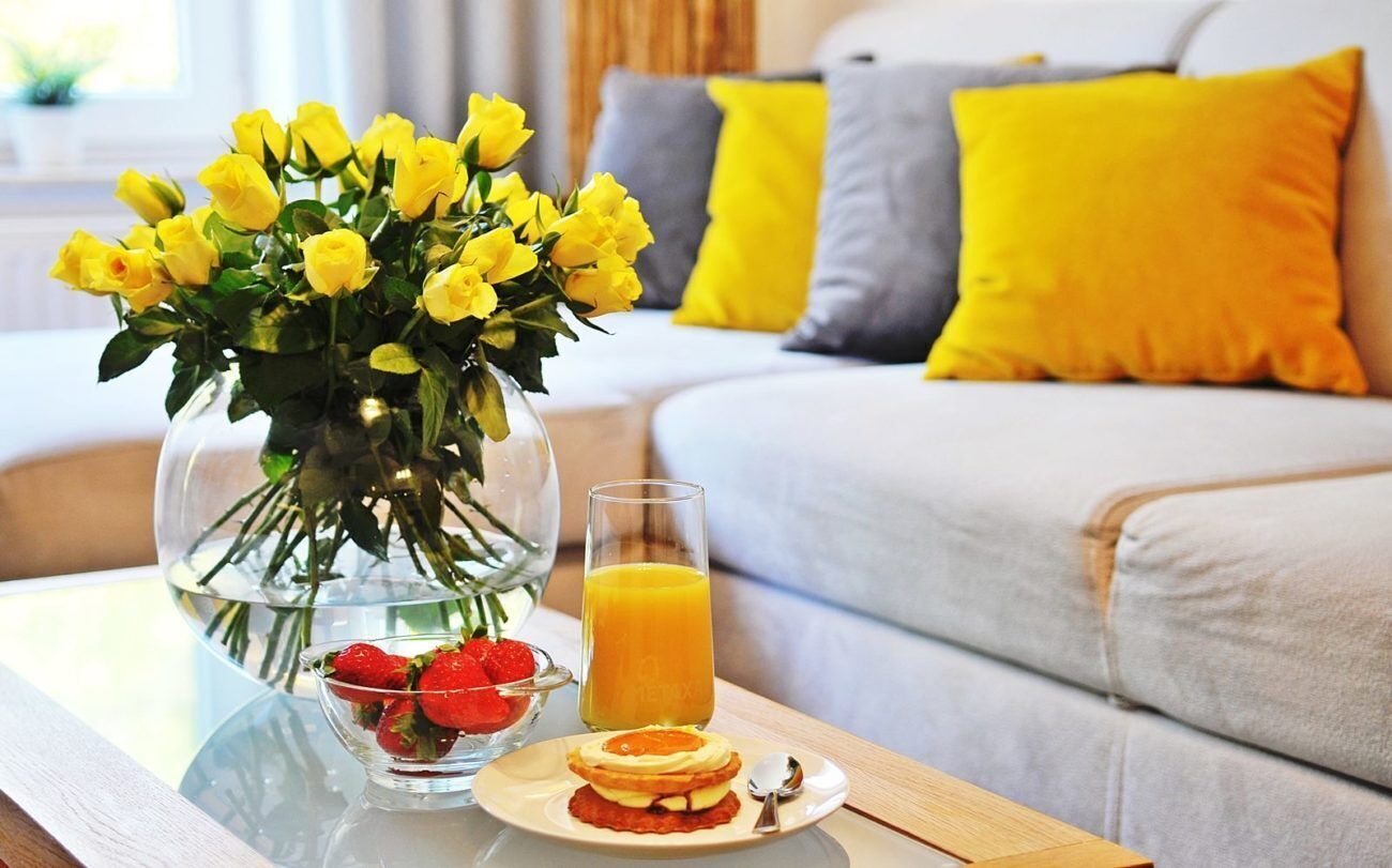 Une table mise pour le petit-déjeuner, avec un vase de fleurs jaunes, un verre de jus d’orange et une assiette de nourriture.