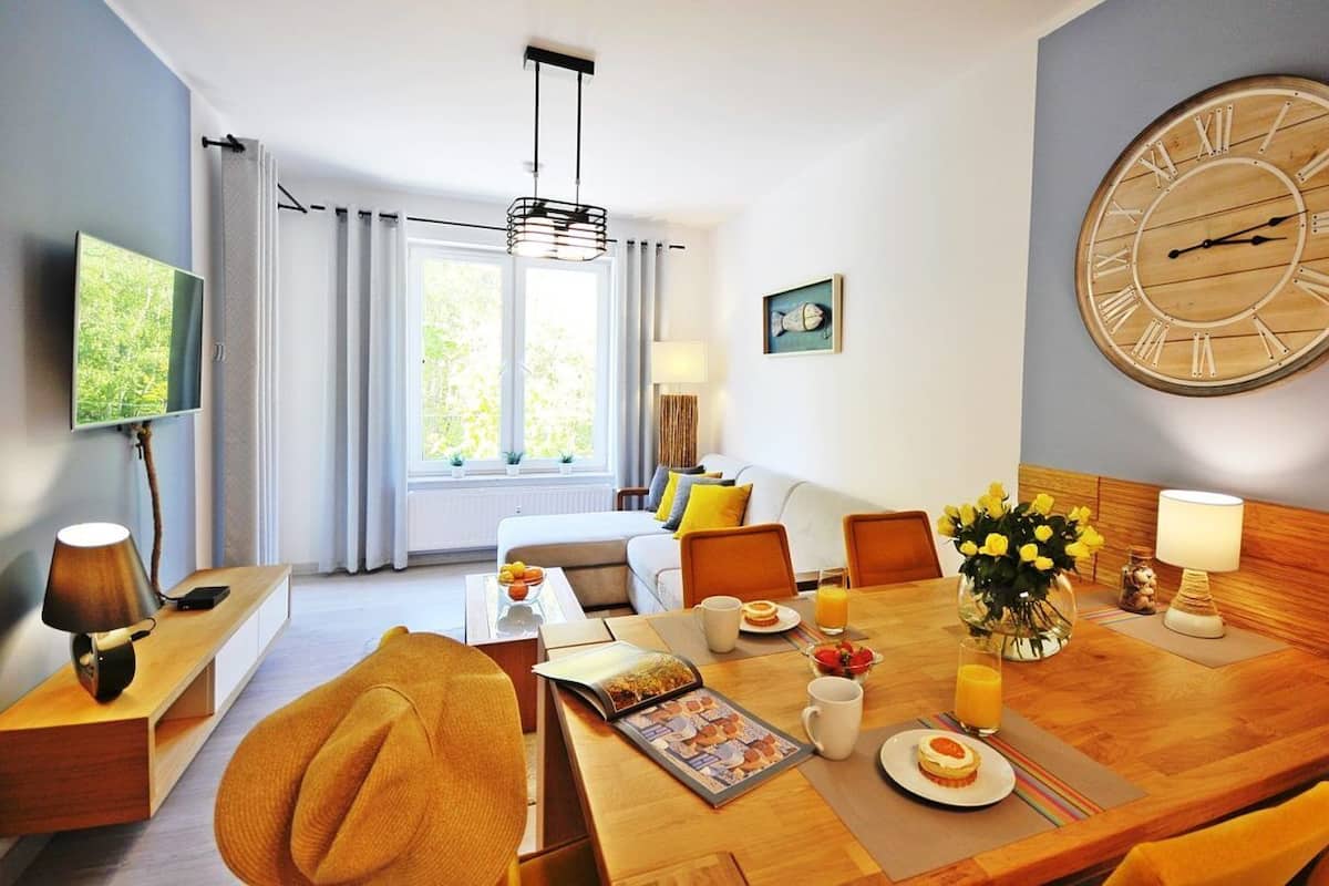 Ein Hotelzimmer mit Essbereich, einer großen Holzuhr, einem Fernseher und einem Fenster mit Vorhängen.