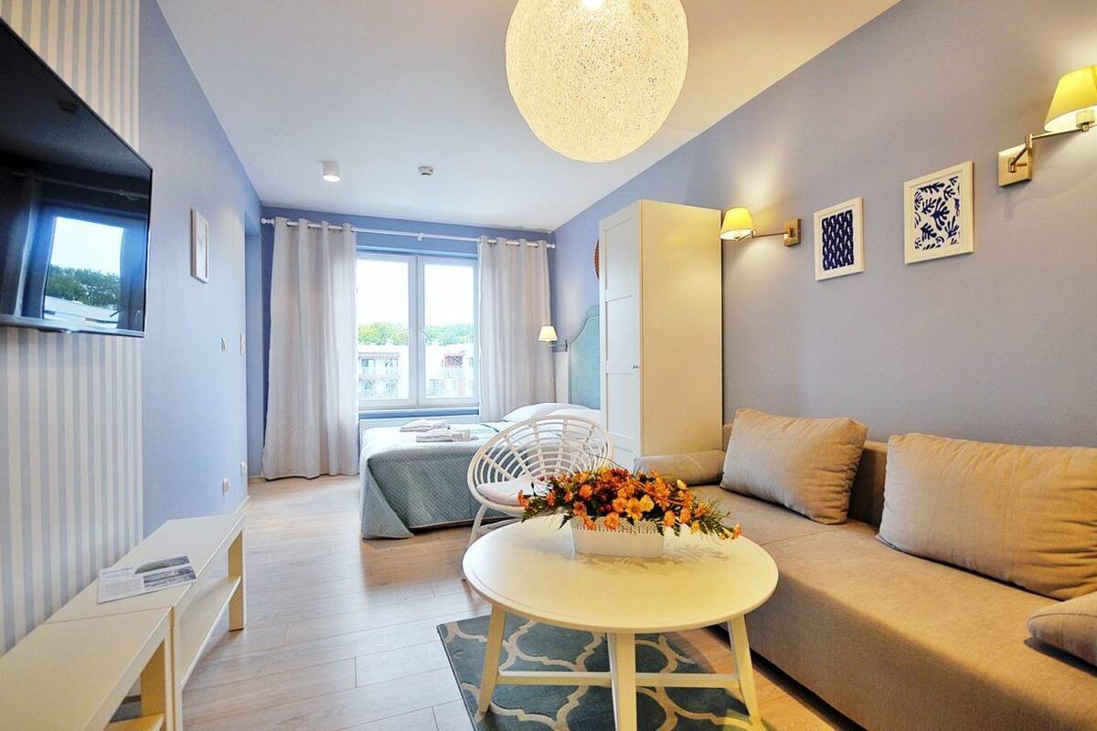 Ein modernes Wohnzimmer mit einer Couch, einem runden Couchtisch mit Blumenarrangements, einem weißen Sessel und einem an der Wand befestigten Fernseher.