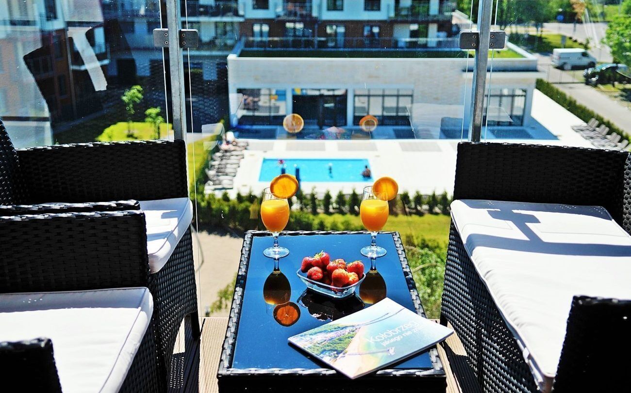 Un balcon avec des fauteuils en osier, une table avec deux verres de jus d’orange, un saladier rempli de fraises et un magazine.