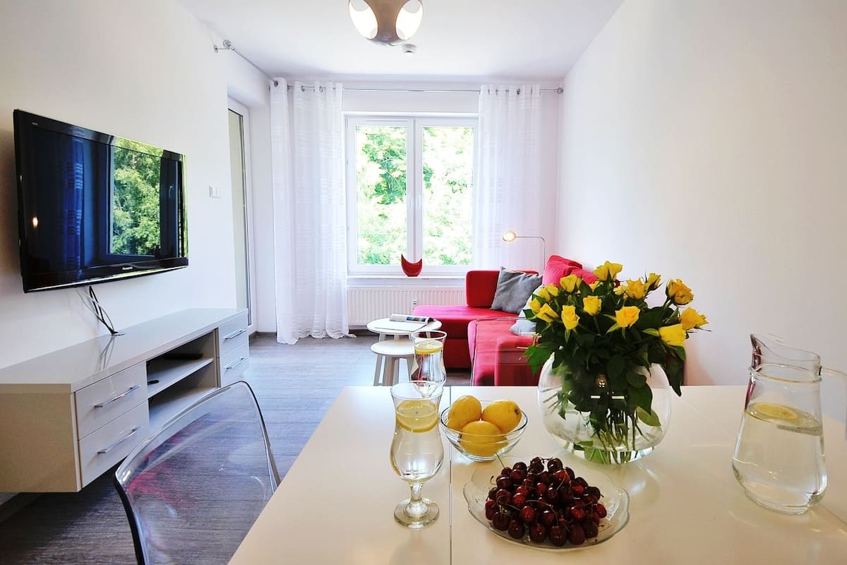 Ein modernes Wohnzimmer mit Fernseher, einem Esstisch mit einer Vase voller gelber Blumen, einer Schale mit Kirschen und einer Karaffe Wasser.