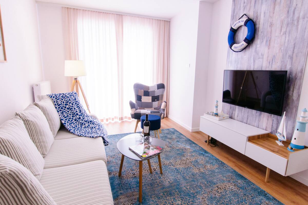 Ein Wohnzimmer mit einer Couch, einem gemusterten Sessel, einem kleinen Couchtisch, einem Fernseher und einer Holzkommode mit Dekorationen.