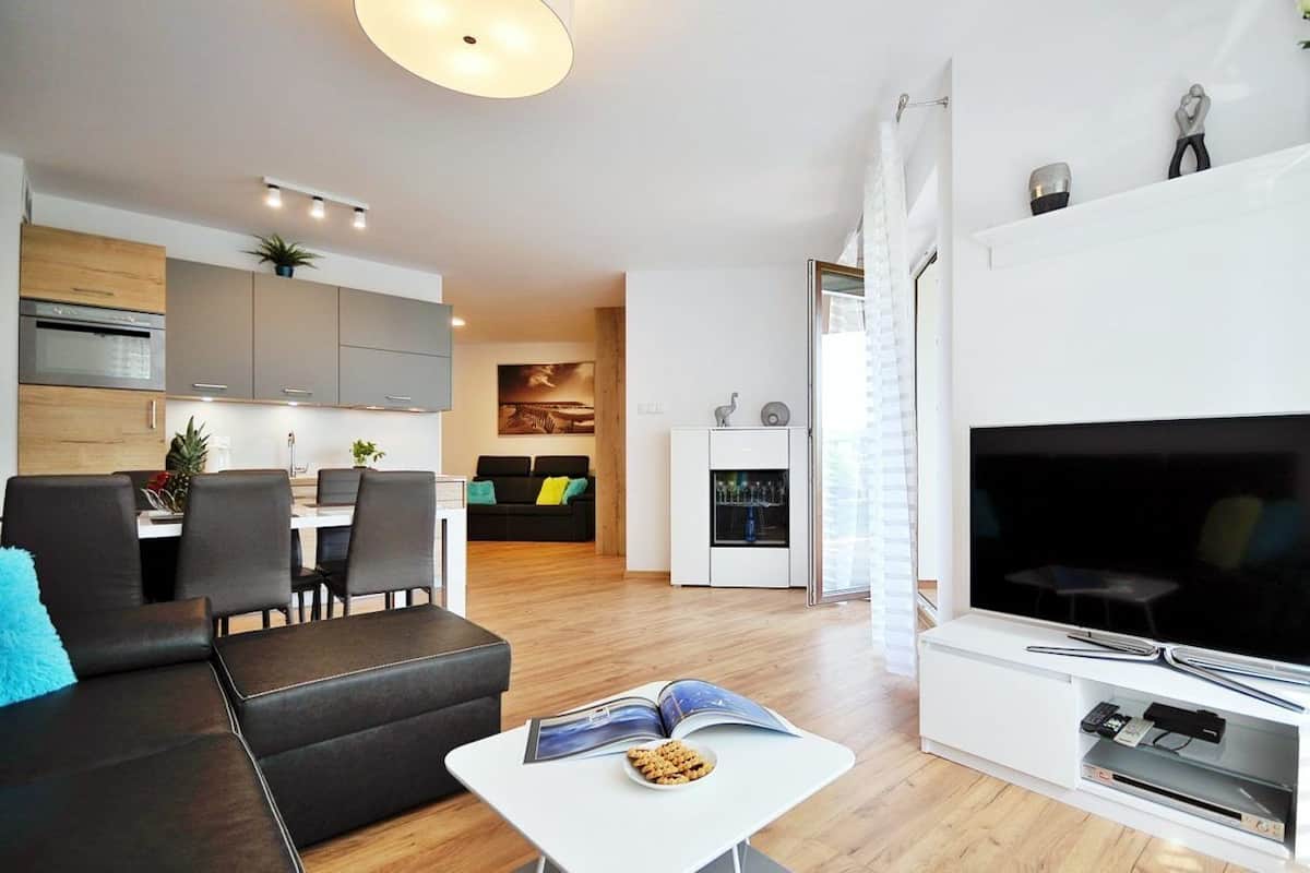 Ein modernes Wohnzimmer mit einer Couch, einem Couchtisch und einem Flachbildfernseher. Im Hintergrund ist eine Essecke mit Tisch und Stühlen zu sehen.