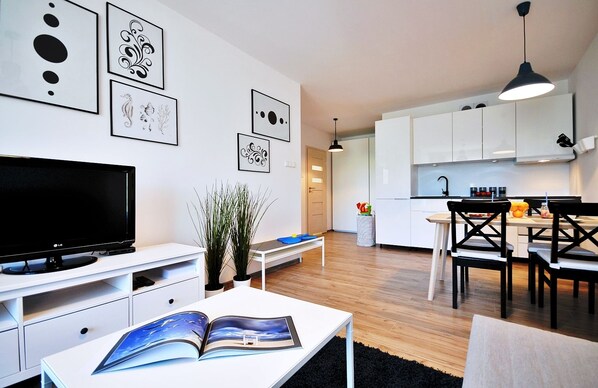 Una sala moderna con una televisión de pantalla plana, una mesa de centro con una revista, y un comedor con una cocina al fondo.
