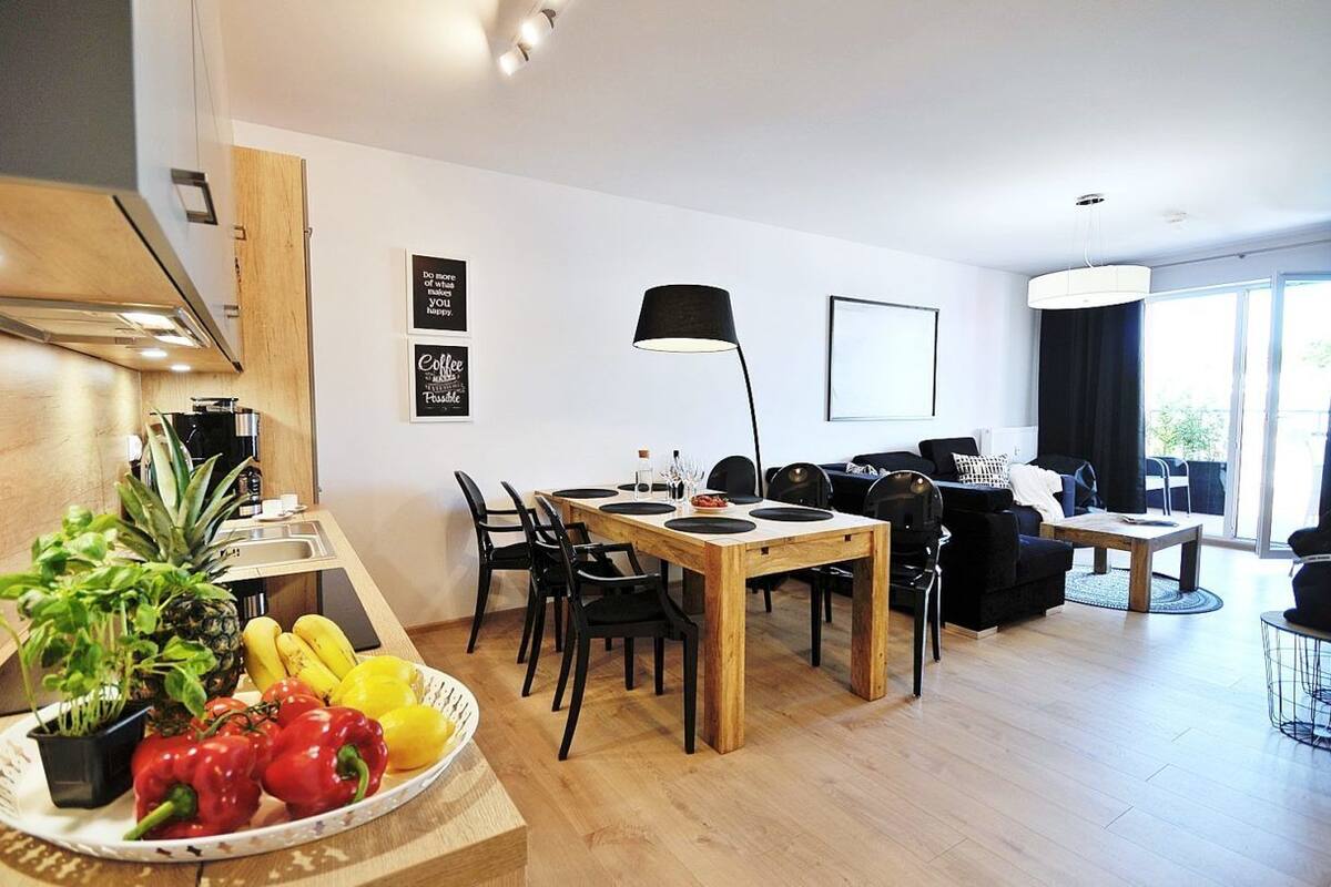 Ein modernes Wohnzimmer mit Essbereich, ausgestattet mit einem Holztisch und -stühlen, einer schwarzen Stehlampe und einem großen Fenster, das für natürliches Licht sorgt.